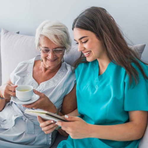 Happy patient is holding caregiver for a hand while spending time together. Elderly woman in nursing home and nurse. Aged elegant woman and tea time at nursing home