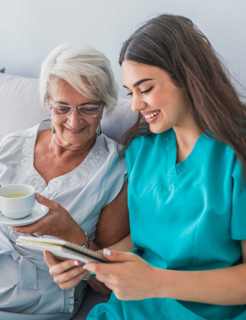 Happy patient is holding caregiver for a hand while spending time together. Elderly woman in nursing home and nurse. Aged elegant woman and tea time at nursing home